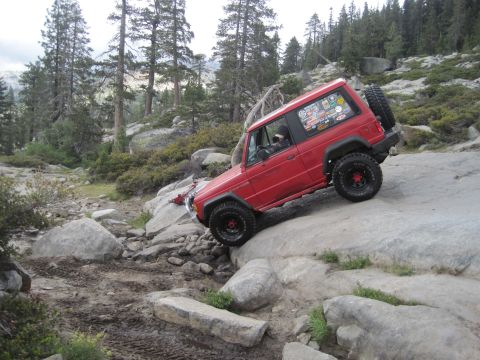 1989 Ford Bronco - A red SUV navigates a challenging rocky trail in a mountainous, wooded area.