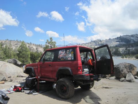 1989 Ford Bronco - Red Jeep Cherokee XJ with off-road modifications parked near a lake on a sunny day.