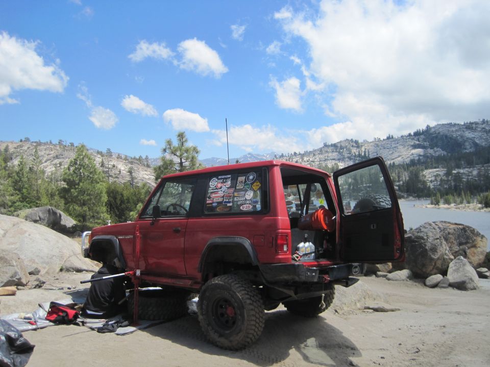 Red 1989 Jeep Cherokee XJ parked on dirt near a lake, rear door open, jack under front wheel