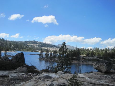 1989 Ford Bronco - A mountain lake scene with scattered rocks and pine trees under a bright blue sky dotted with white clouds.