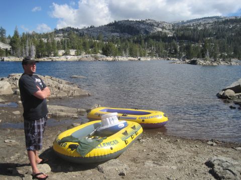 1989 Ford Bronco - Two inflatable rafts sit on the rocky shore of a lake, with a man standing nearby and mountains in the distance.