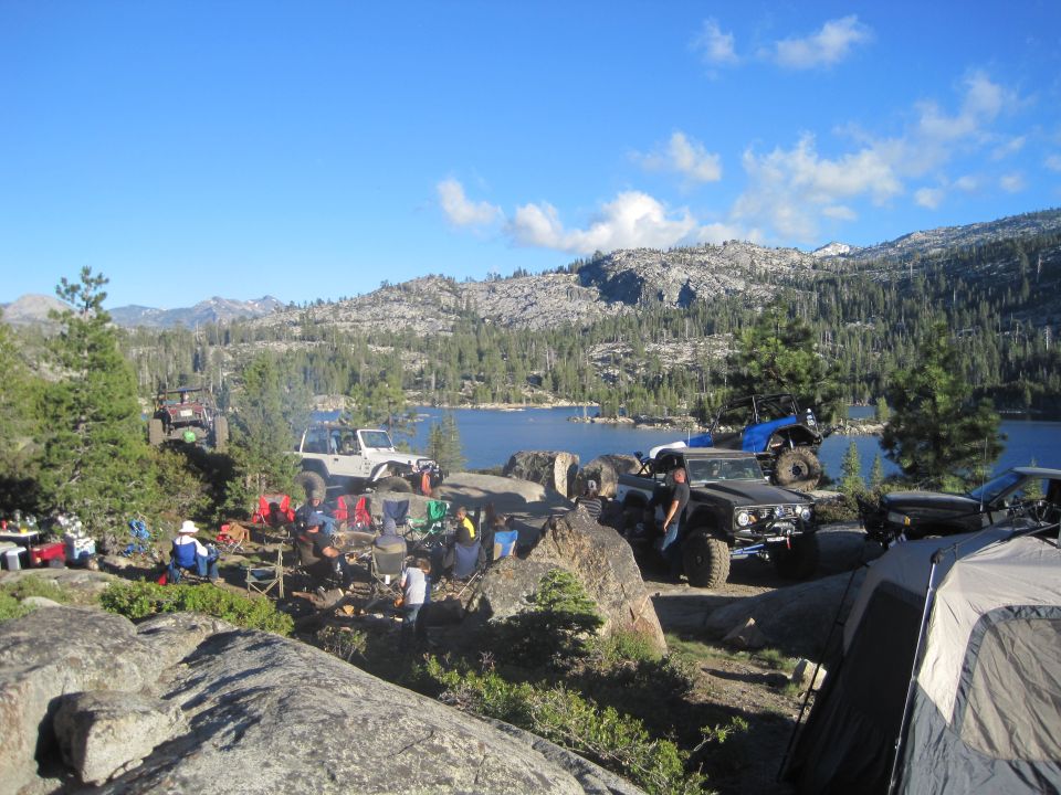 Several off-road vehicles parked on rocky terrain near a lake with trees and mountains in the background