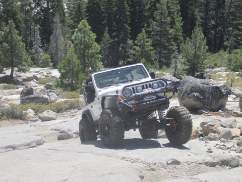 1989 Ford Bronco - A modified white Jeep navigates a challenging rocky trail in a wooded area.