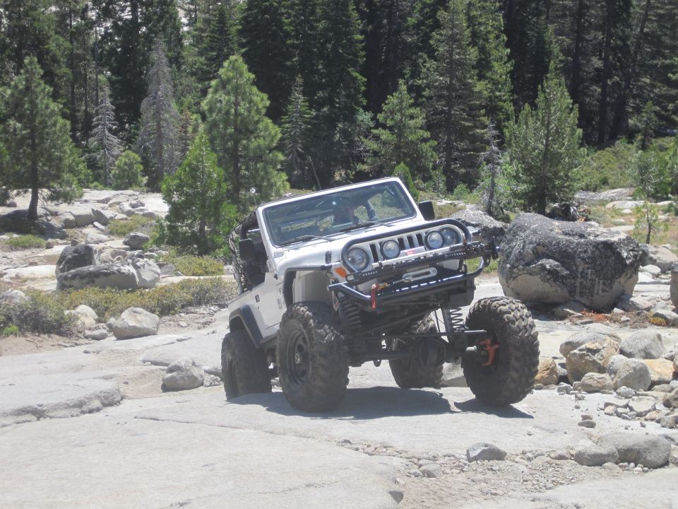 White Jeep Wrangler with off-road modifications, including a winch and large tires, navigating rocky terrain