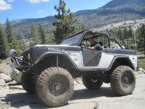 1989 Ford Bronco - Modified off-road Bronco with prominent tires and roll cage navigates a rocky terrain.
