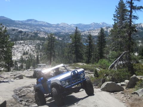 1989 Ford Bronco - Two off-road vehicles navigate a challenging rocky trail in a mountainous, wooded landscape under a clear blue sky.
