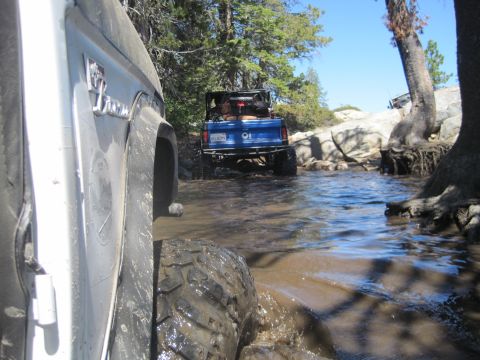 1989 Ford Bronco - Two Broncos navigate a muddy stream on a sunny day, with the lead vehicle's side and tire prominent in the foreground.