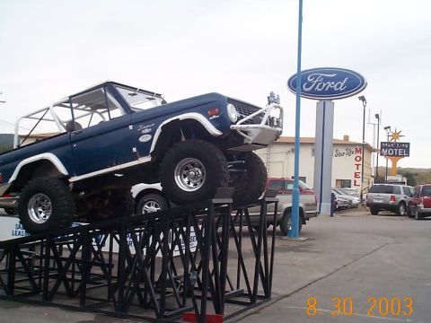 1977 Ford Bronco - A blue 1977 Ford Bronco is displayed on ramps in front of a Ford dealership and a motel.