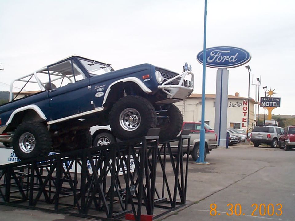 Blue early Ford Bronco on white wagon wheels displayed on black ramps under a Ford dealership sign
