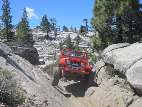 1989 Ford Bronco - An orange Jeep Wrangler navigates a challenging rocky off-road course under a clear blue sky.
