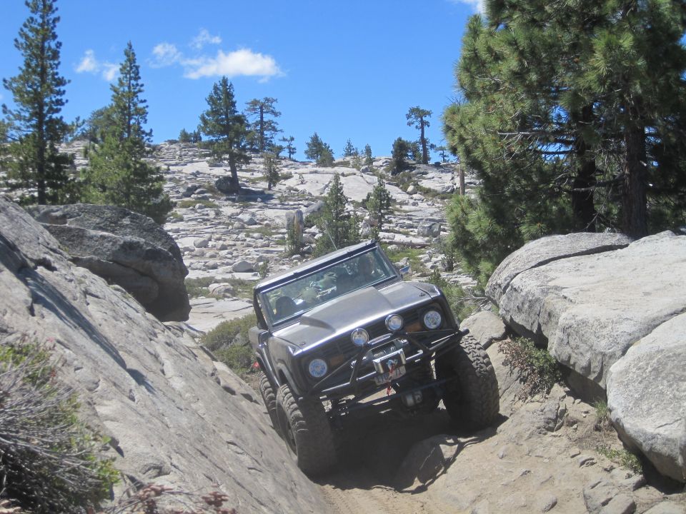 1989 Ford Bronco navigating a steep, rocky trail with large boulders and pine trees