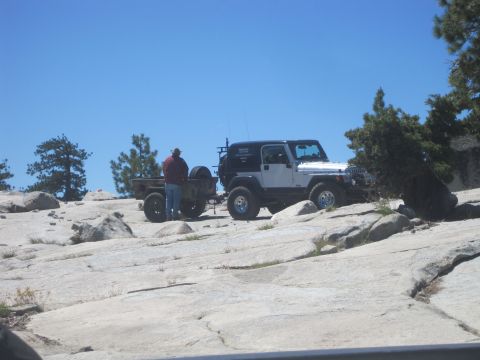 1989 Ford Bronco - A white Jeep Wrangler tows a camouflage trailer across a rocky landscape under a clear blue sky.