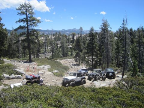 1989 Ford Bronco - A group of off-road vehicles navigates a trail through a forested, mountainous landscape under a clear blue sky.