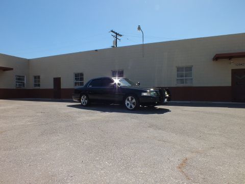 2003 Ford Crown Victoria - Wide shot of a black sedan with sun glare parked across a paved lot from a beige building