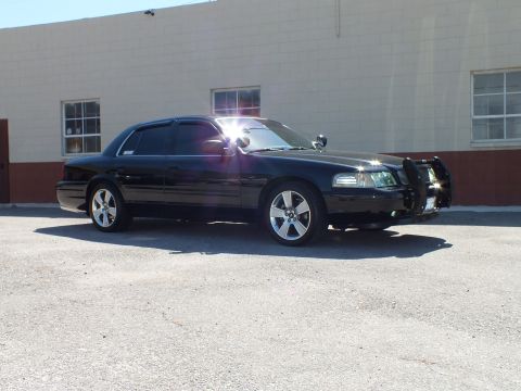 2003 Ford Crown Victoria - Black sedan front quarter with chrome push bumper and pillar spotlights, sun glare on windshield