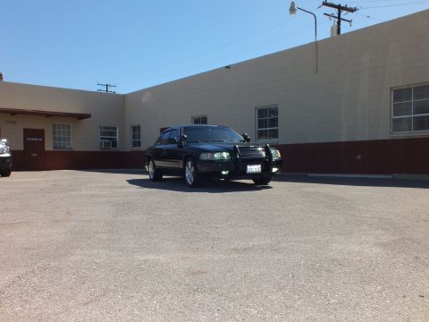 2003 Ford Crown Victoria - Distant front view of a dark Crown Victoria with grille guard in a paved lot near a low warehouse.