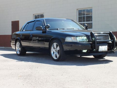 2003 Ford Crown Victoria - Right side-front view of a black sedan with EVIL VIC plate parked by a tan wall.