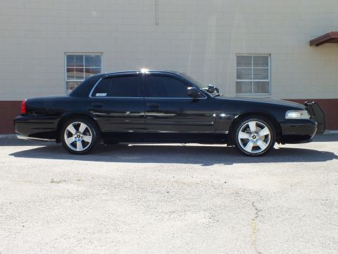 2003 Ford Crown Victoria - Driver-side profile of a lowered black sedan on chrome wheels by a tan building.