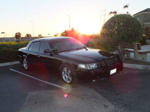2003 Ford Crown Victoria - Front-right view of a black sedan backlit by a setting sun, lens flare across frame.
