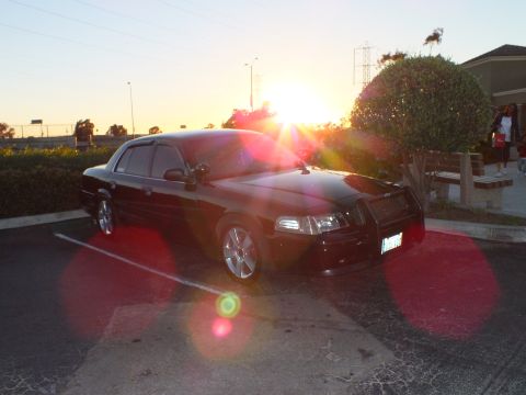 2003 Ford Crown Victoria - Black sedan parked facing the sunset, strong sunburst and red lens flare in shot.