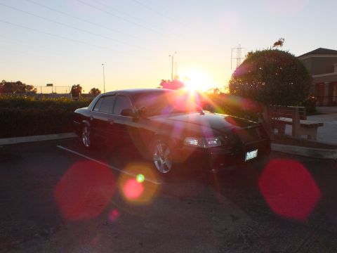 2003 Ford Crown Victoria - Front-right view of a black sedan against a bright sunset with heavy lens flare.