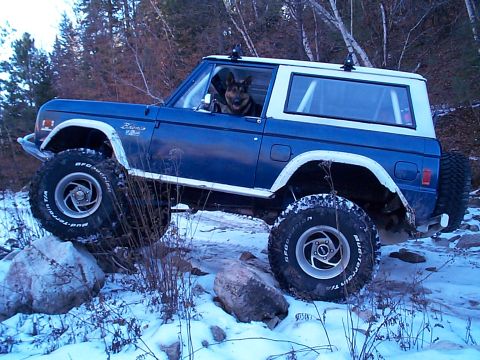 1977 Ford Bronco - Blue early Ford Bronco with a white hardtop and white fender flares parked on snowy ground among rocks and bare trees, driver-side profile…