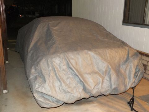 2003 Ford Crown Victoria - Vehicle shrouded in a tan and gray car cover, photographed beside a brick wall in a driveway.