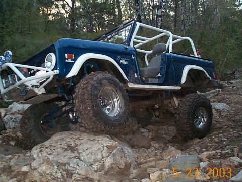 1977 Ford Bronco - Driver-side view of a blue topless early Ford Bronco with white roll cage, white fender flares and white tube front bumper crawling across…