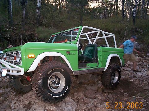1977 Ford Bronco - A green 1977 Ford Bronco with a white roll cage navigates a rocky trail, with a person exiting the driver's side.