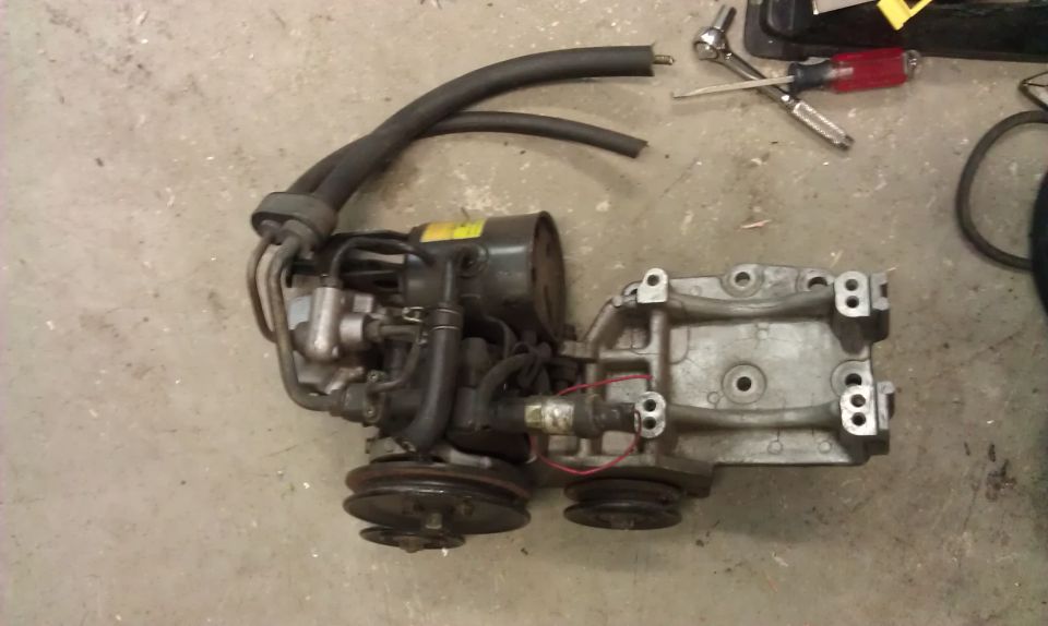Engine component with hoses and pulleys on a concrete floor. Tools are visible in the background