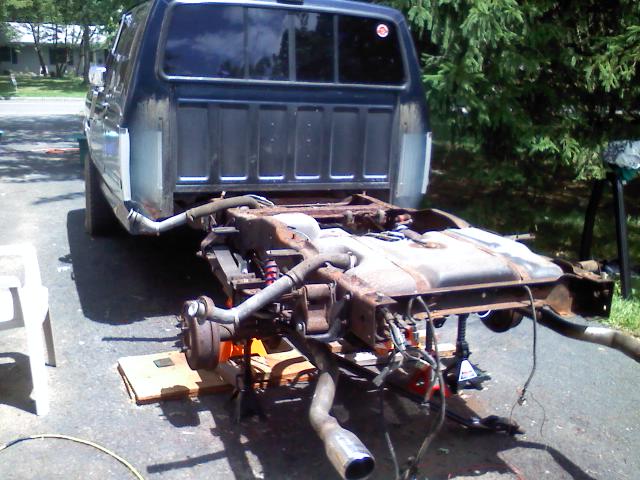 Dark blue pickup with bed lifted off rusty frame showing rear axle, fuel tank and exhaust in a driveway
