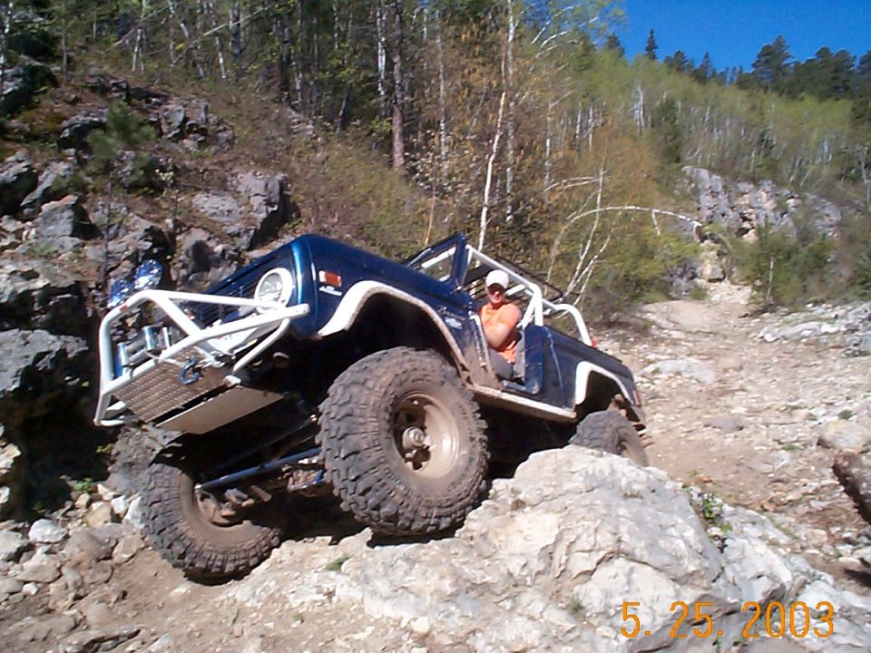 Blue 1977 Ford Bronco climbing rocky terrain with a driver visible