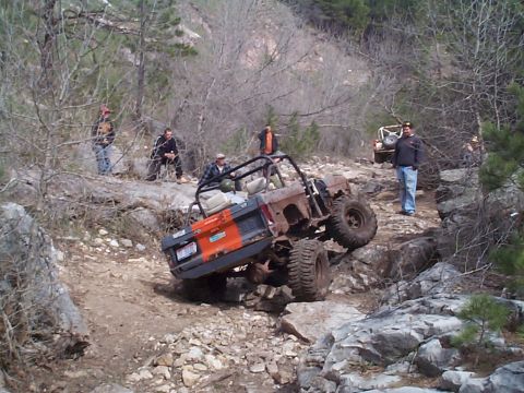 1977 Ford Bronco - Rear view of a topless orange and gray primered early Ford Bronco with an interior roll cage flexing over a rocky off-road trail, one rear…