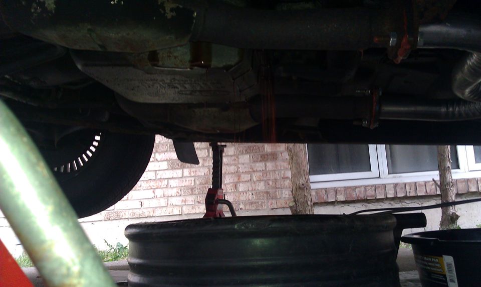 Underside of a vehicle with transmission fluid draining into a pan, with a jack visible