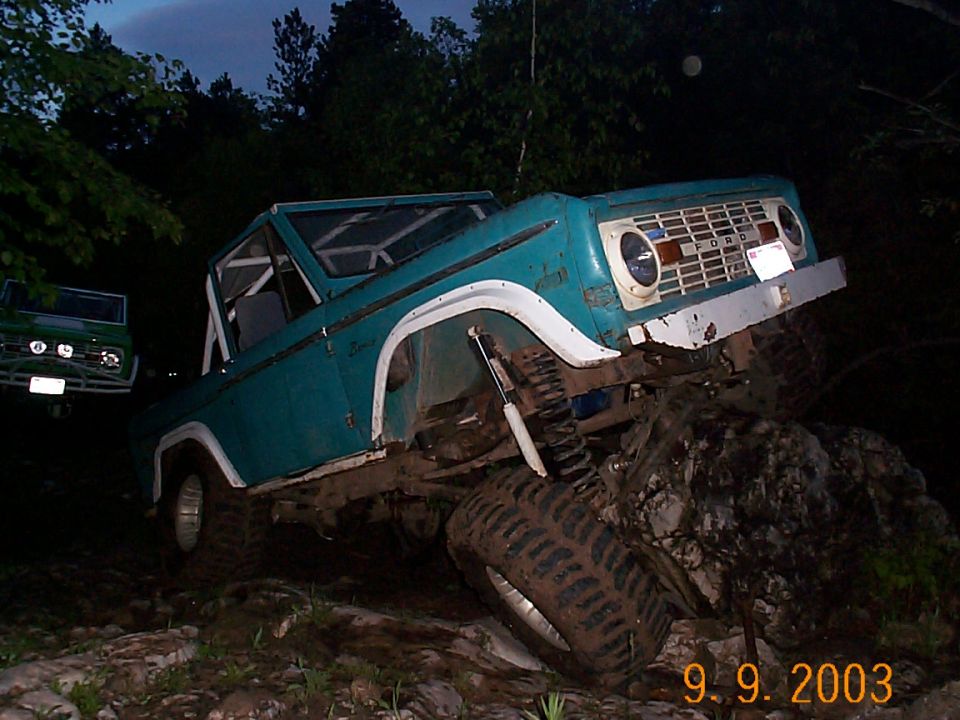 Teal Ford Bronco off-roading, climbing over rocks. Another green Bronco is visible in the background