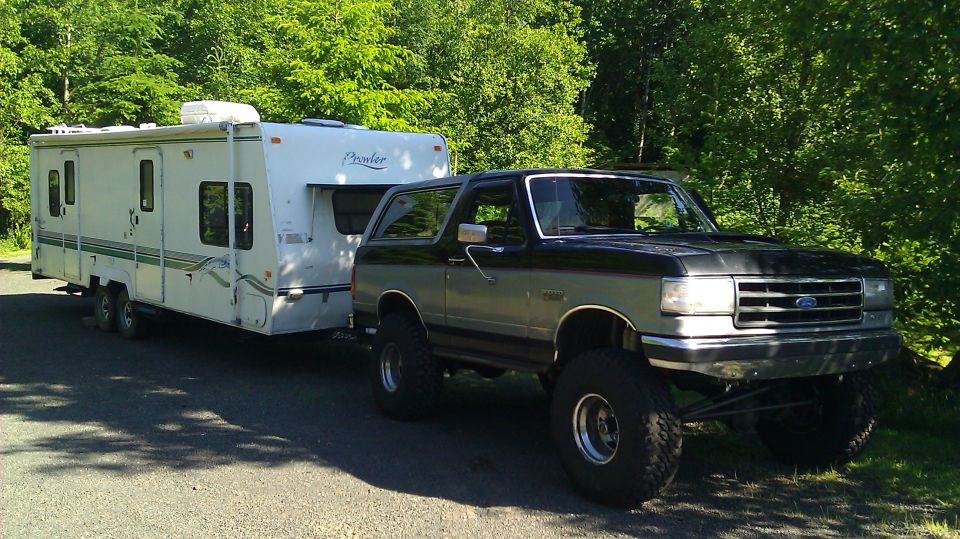 Lifted black and gray Ford Bronco on mud tires towing a white Prowler travel trailer on gravel