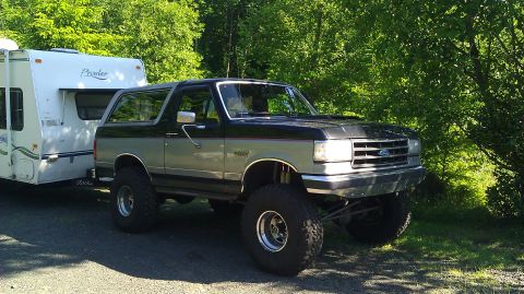 1989 Ford Bronco - This lifted Ford Bronco is hitched to a white travel trailer and parked on a gravel road.