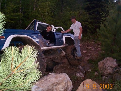 1977 Ford Bronco - A blue 1977 Ford Bronco is shown off-roading through a rocky, wooded area with two individuals.