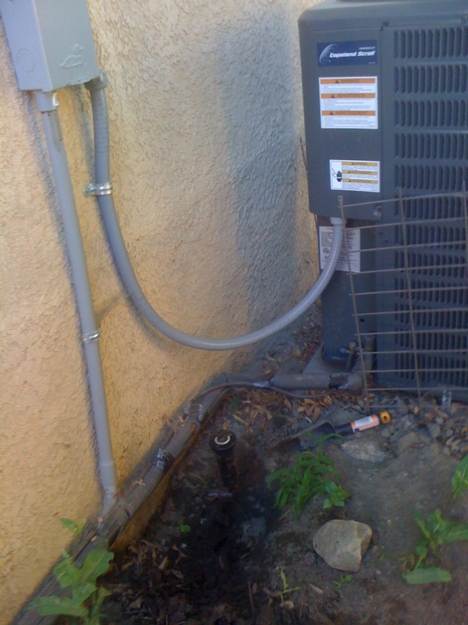 Outdoor air conditioning unit next to a stucco wall with electrical conduit and a sprinkler head visible