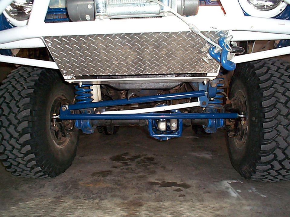 Front underside of lifted blue Bronco showing axle, coil springs, blue steering links, diamond-plate skid plate and big tires