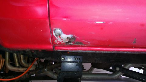 1978 Ford Bronco - Close-up of rust damage on a red vehicle's lower body panel, with exhaust components visible below.