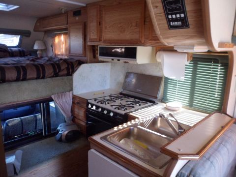 1991 Ford Bronco - The interior of an RV features a raised bed, a kitchen with a stove and sink, and wooden cabinetry.