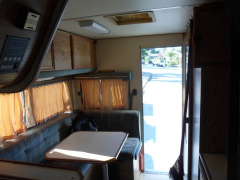 1991 Ford Bronco - The interior of a camper van is visible, with a bench seat, table, and cabinets, looking out an open door to a street.