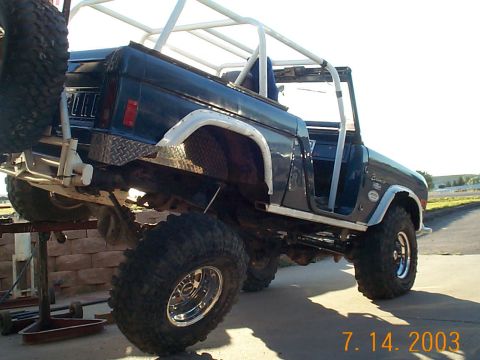 1977 Ford Bronco - Low driver-side rear angle of a dark blue topless early Ford Bronco lifted at the rear on a jack stand, showing white roll cage, white fend…
