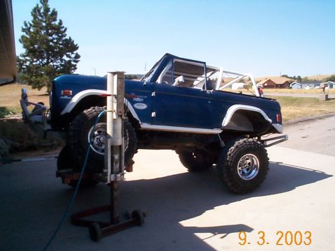 1977 Ford Bronco - Blue topless early Ford Bronco with a white roll cage and white fender flares, passenger side view on a driveway with the rear wheel remove…