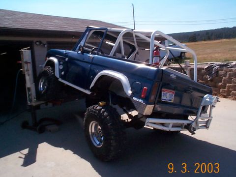 1977 Ford Bronco - Blue topless early Ford Bronco raised nose-down at the front on a shop jack in a driveway, seen from the passenger-side rear with a white r…