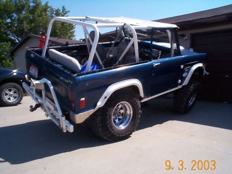 1977 Ford Bronco - Blue topless early Ford Bronco parked in a driveway, viewed from the driver-side rear with a white full roll cage, white swing-out tire car…