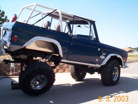 1977 Ford Bronco - Dark blue topless early Ford Bronco on a driveway, shown from the driver's side with a bare white roll cage, white fender flares, diamond-p…