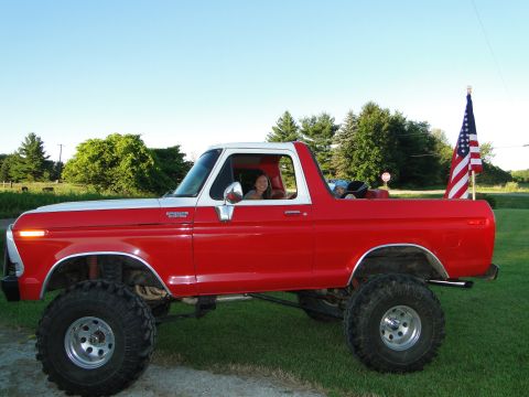 1978 Ford Bronco - A lifted red and white 1978 Ford Bronco is parked on a grassy area with trees and a blue sky in the background.