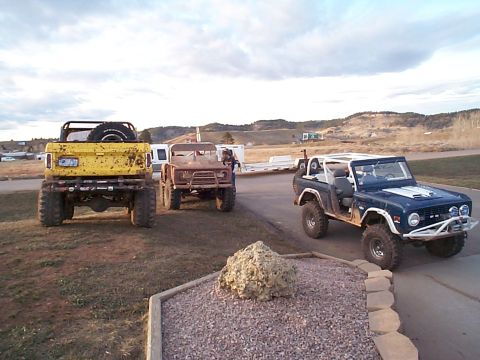 1977 Ford Bronco - Three modified Ford Broncos are parked outdoors with a scenic hilly landscape behind them.